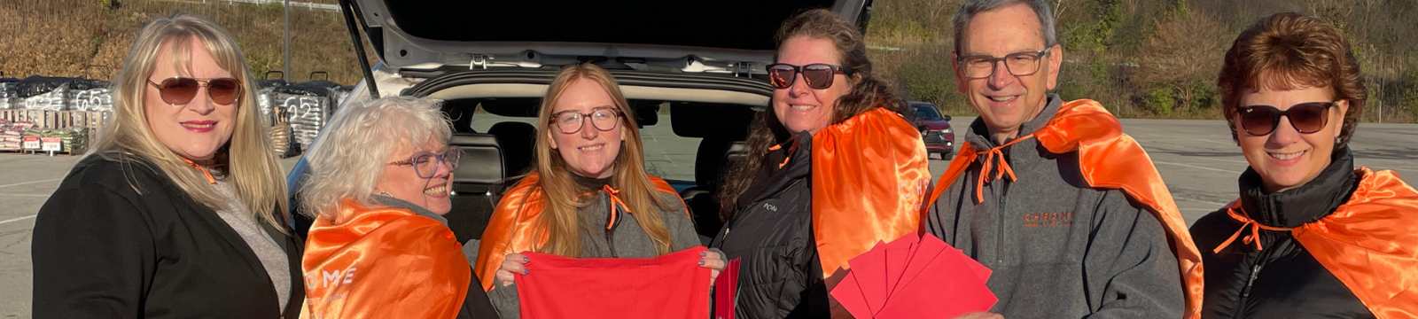CHROME Employees outside a vehicle holding red cards, a bag, and wearing capes, smiling for a photo.